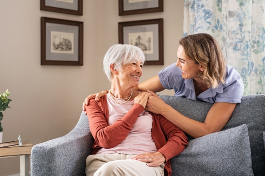 Feasterville-Trevose A smiling senior woman sits on a couch while a cheerful caregiver gently holds her shoulders, both sharing a warm moment in a cozy home setting. This image represents compassionate home care in Feasterville-Trevose, highlighting the supportive and trusting relationship between caregivers and seniors.