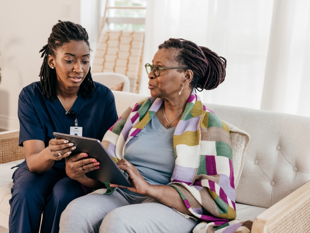 A caregiver in blue scrubs supports a senior woman with a tablet while sitting together on a couch, representing compassionate home care in Bala Cynwyd.