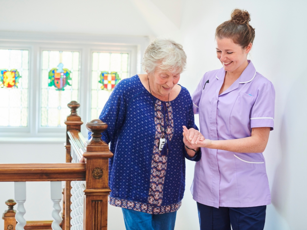 A smiling caregiver in a purple uniform supports an elderly woman walking indoors, representing compassionate home care in Old City/Society Hill.